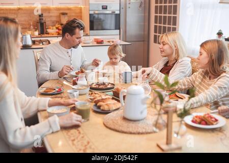 Portrait grand angle de la grande famille heureuse appréciant le petit déjeuner ensemble dans la cuisine, se concentrer sur le père aidant mignon bébé fille, copier l'espace Banque D'Images