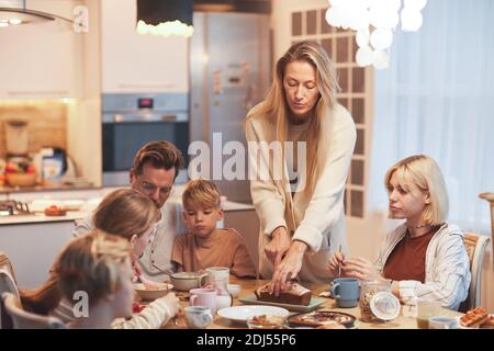 Portrait grand angle de la grande famille heureuse appréciant le petit déjeuner ensemble assis à table dans la cuisine, se concentrer sur la mère servant la nourriture et couper le gâteau, copie Banque D'Images