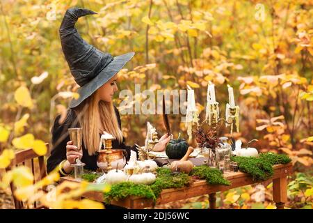 Jeune femme en costume de sorcière assise à table avec magie ingrédients et notes le jour de l'automne en forêt Banque D'Images