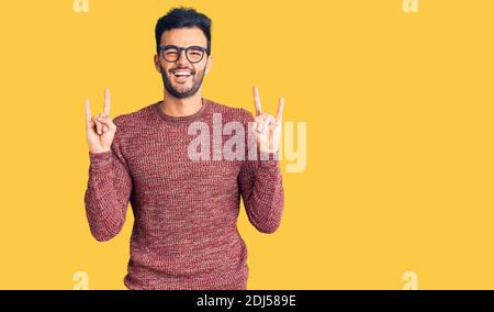 Jeune homme hispanique élégant portant un chandail d'hiver et des lunettes criant avec l'expression folle faisant le symbole de rock avec les mains vers le haut. Star de la musique. Conce lourd Banque D'Images