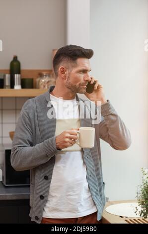 Vertical taille vers le haut portrait de beau homme mature parlant par smartphone et prendre une tasse de café le matin tout en étant debout par fenêtre dans la cuisine Banque D'Images