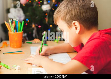 7 yeas vieux garçon assis à un bureau et faisant des devoirs. L'enfant écrit des notes dans le carnet, les devoirs difficiles, l'enfant se concentrant sur des exemples. Arbre de Noël décoré sur fond Banque D'Images