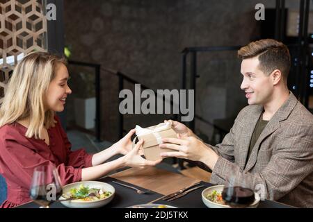 Joyeux jeune homme passant boîte-cadeau avec cadeau pour la Saint-Valentin à sa jolie petite fille tout en étant assis à la table de service et dîner Banque D'Images