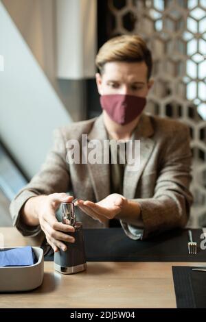 Mains de jeune homme élégant pressant distributeur de pot en plastique avec de l'assainisseur avant de dîner assis près d'une table restaurant ou café Banque D'Images