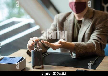 Mains de jeune homme contemporain distributeur de pression de pot en plastique avec de l'assainisseur avant de dîner assis près d'une table restaurant Banque D'Images