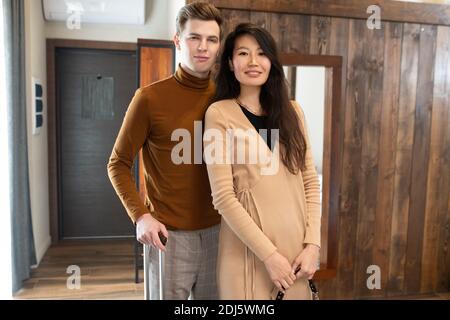 Jeune femme asiatique en élégante robe beige vous regardant avec un sourire éclatant avec son mari ou son petit ami avec une valise à proximité Banque D'Images