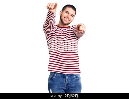 Jeune homme beau portant un chandail rayé pointant vers vous et l'appareil photo avec les doigts, souriant positif et gai Banque D'Images