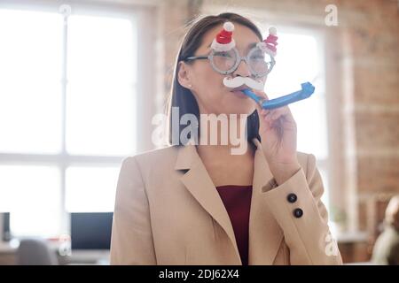 Jeune femme d'affaires élégante en lunettes de Noël avec moustache blanche debout devant l'appareil photo au bureau et sifflement d'anniversaire Banque D'Images