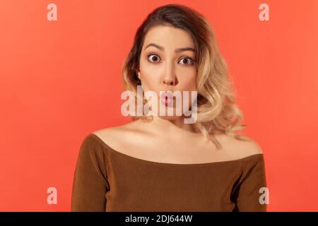 Portrait de drôle jeune belle blonde femme en blouse brune debout et regardant l'appareil photo avec des lèvres de poisson. Studio d'intérieur isolé sur b rouge Banque D'Images