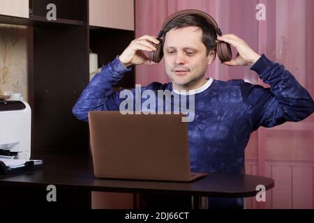 Un homme dans les vêtements de la maison travaille à un ordinateur portable. Met un casque sur la tête. Banque D'Images
