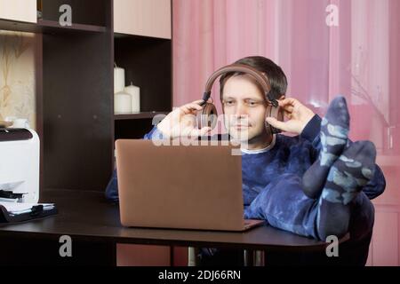 Un homme dans les vêtements de la maison travaille à un ordinateur portable. Met un casque sur la tête. Il a mis ses pieds sur la table. Banque D'Images
