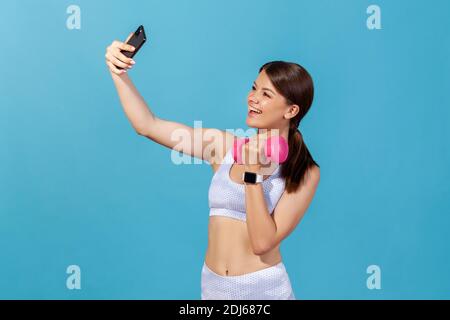 Souriante bonne femme sportive en blanc haut de sport et collants faisant selfie y appareil photo de smartphone pompant les muscles et l'entraînement à la salle de gym. St. Intérieure Banque D'Images