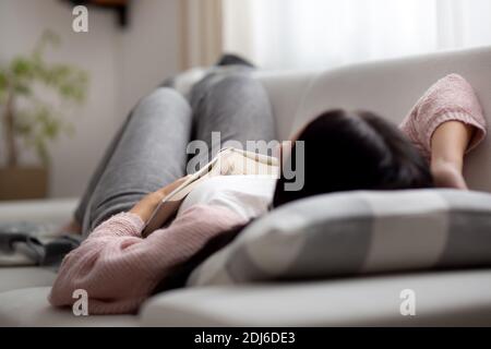 Une femme qui s'endormit pendant la journée sur un canapé tout en essayant de lire un livre. Banque D'Images