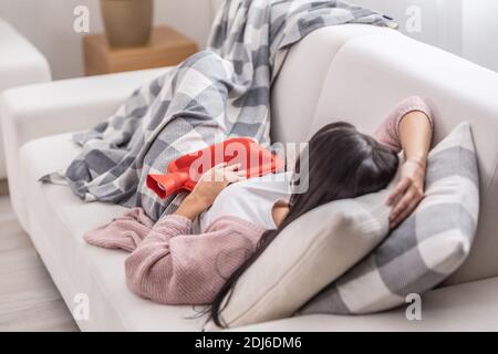 Estomacache se soulagant par thermophore tandis que la femme est sur un canapé sous la couverture. Banque D'Images