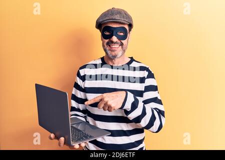 Moyen âge effarant cambrioleur homme portant un chapeau et un masque en utilisant ordinateur portable sur fond jaune sourire heureux pointant avec la main et doigt Banque D'Images