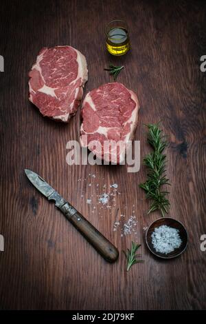 Deux steaks de bœuf agrémenté d'une brindille de romarin, d'huile d'olive, de granules de sel de mer et d'un couteau sur fond de bois. Banque D'Images