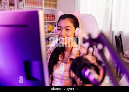 Une jeune fille de gamer excitée et souriante dans un micro-casque avec un jeu vidéo en ligne et la diffusion en direct. Jeune femme asiatique parlant à des joueurs et à un public sur ordinateur dans une salle lumineuse à LED néon à la maison Banque D'Images