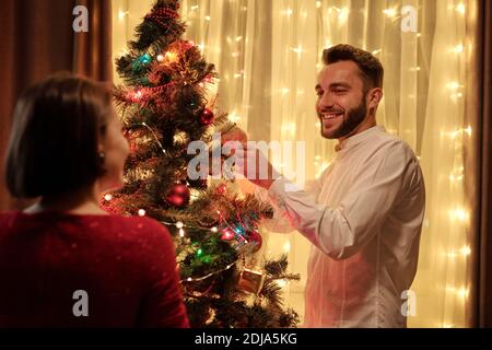 Homme barbu souriant en chemise blanche décorant arbre de Noël avec petite amie pendant qu'ils se préparent à la fête Banque D'Images