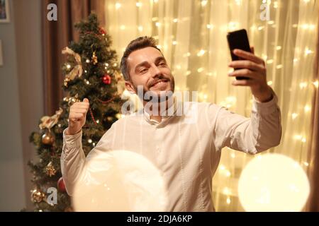 Souriant beau jeune homme en chemise blanche pointant à Noël arbre tout en prenant le selfie dans la salle de séjour Banque D'Images