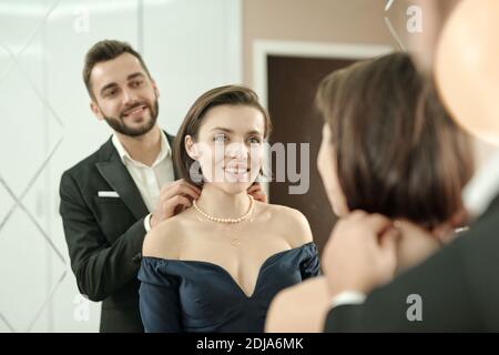 Homme barbu souriant en costume portant un collier à la petite amie devant le miroir pendant qu'ils s'habillez pour dîner restaurant Banque D'Images
