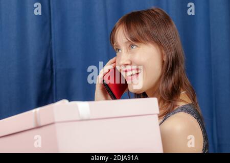 adorable jeune fille à cheveux bruns tenant une boîte rose avec un offrez-lui dans ses mains et parlez au téléphone un arrière-plan bleu Banque D'Images