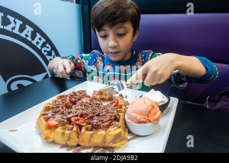 Un jeune garçon portant un pull de Noël mange une grande gaufre avec de la glace, des fraises et du chocolat. Banque D'Images