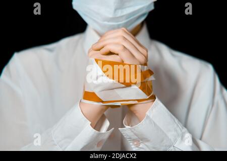 Mains d'un homme dans un masque médical, attaché avec un stop-ruban, gros plan. Femme attachée dans un masque de protection sur fond noir, le concept d'un coronavirus Banque D'Images