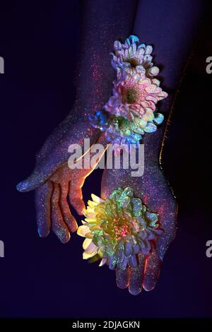 Les mains d'une femme en lumière ultraviolette avec des fleurs dans les paumes. Cosmétiques pour le soin de la peau des mains Banque D'Images