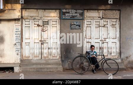 Jamnagar, Gujarat, Inde - décembre 2018 : un Indien âgé assis sur la chaussée avec son vélo devant les volets fermés d'un magasin. Banque D'Images