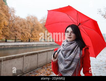 portrait d'une jeune fille souriante en robe rouge avec un parapluie se tenant sur la ruelle du parc après la pluie le jour sombre de l'automne Banque D'Images