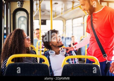 Jeune heureux adorable couple est assis ensemble sur un bus et est accueilli par un jeune homme beau qu'ils connaissent tous les deux. Banque D'Images