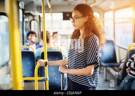 Jeune fille mignonne avec des cheveux bouclés est tenue sur la barre et ses bagages tout en se tenant dans un bus. Banque D'Images