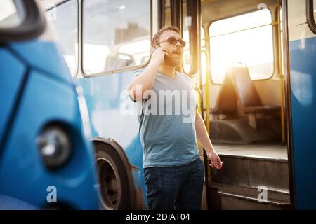 Le jeune homme parle au téléphone et fume une cigarette tout en se tenant à côté du bus bleu. Banque D'Images