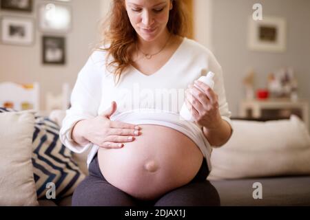 Belle femme enceinte appliquant une crème sur son ventre tout en étant assise à la maison. Banque D'Images