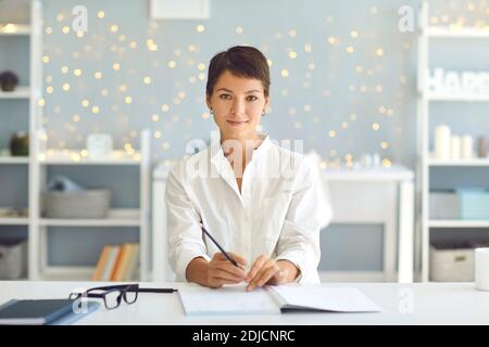 Femme assurée assise au bureau avec un stylo et un ordinateur portable ouvert, regardant l'appareil photo comme si l'enregistrement vidéo Banque D'Images