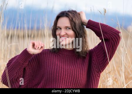 Portrait d'une jolie jeune femme dans un chandail brun se tient dans l'herbe séchée dans le champ. Femme caucasienne attrayante avec de longs cheveux bruns et des lèvres rouges. Banque D'Images