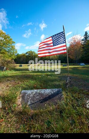 Un drapeau américain vole à côté de la pierre tombale d'un ancien vétéran militaire dans un cimetière rural. Au cimetière de Morgan Bay à Surry, Maine. Banque D'Images