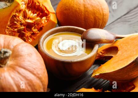 Soupe de potiron d'automne à la crème aigre dans un bol en bois et citrouilles coupées fraîches sur fond sombre Banque D'Images