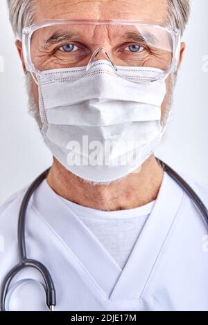 homme confiant médecin dans le masque, homme âgé mature regarde la caméra portant un costume médical blanc et des lunettes de protection, en utilisant le stéthoscope, fond blanc Banque D'Images
