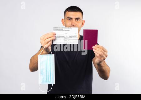 Un homme tient devant lui une carte de vaccination, un passeport et un masque médical sur fond blanc. Photo de haute qualité Banque D'Images