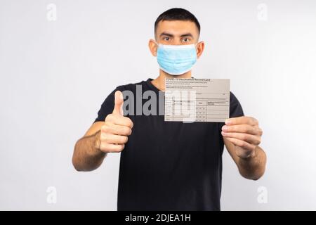 Un homme libre de quarantaine, dans un masque médical tient une carte de vaccination du coronavirus et des gestes de pouce vers le haut. Grand comme pour la vaccination en cas de pandémie. Banque D'Images