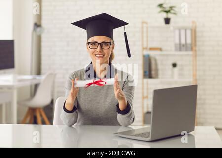 Joyeux cours en ligne diplômé assis à un bureau avec ordinateur portable, tenant son diplôme et souriant Banque D'Images