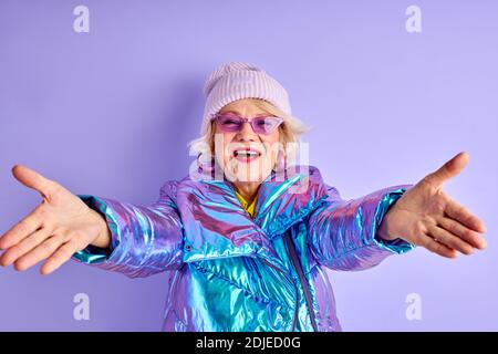 gaie femme âgée avec bras écarteur regarder la caméra, allant à câlin, portant manteau d'hiver chapeau et lunettes de soleil Banque D'Images