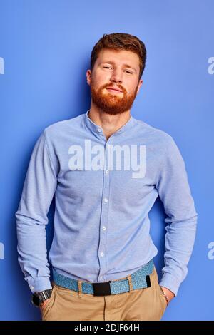 homme barbu en chemise posé à l'appareil photo, homme aux cheveux rouges posant avec les mains dans la poche, isolé sur fond bleu Banque D'Images
