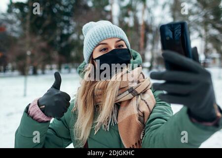 Une femme prend un selfie portant un masque à l'extérieur pendant la pandémie du coronavirus Covid-19. Fille utilisant un smartphone dans un parc hivernal enneigé montrant le pouce vers le haut Banque D'Images