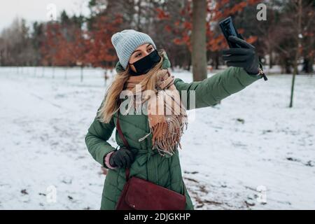 Une femme prend un selfie portant un masque à l'extérieur pendant la pandémie du coronavirus Covid-19. Fille utilisant un smartphone dans un parc hivernal enneigé Banque D'Images