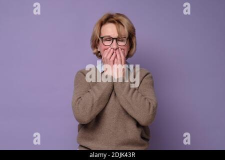 La femme caucasienne senior rigole joyeusement, couvre la bouche pendant que les tentatives cessent de rire étant surpris par les nouvelles. Prise de vue en studio Banque D'Images