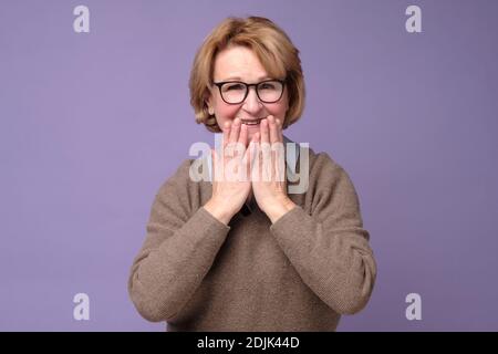 La femme caucasienne senior rigole joyeusement, couvre la bouche pendant que les tentatives cessent de rire étant surpris par les nouvelles. Prise de vue en studio Banque D'Images