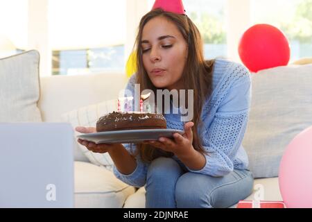 Femme caucasienne ayant anniversaire appel vidéo soufflant sur des bougies gâteau portant chapeau de fête Banque D'Images