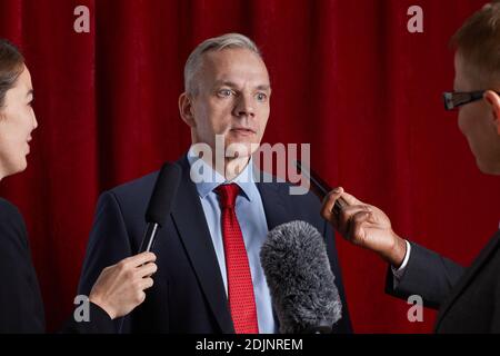 Portrait d'un homme mûr donnant une interview à un journaliste et parler aux microphones contre le rideau rouge Banque D'Images